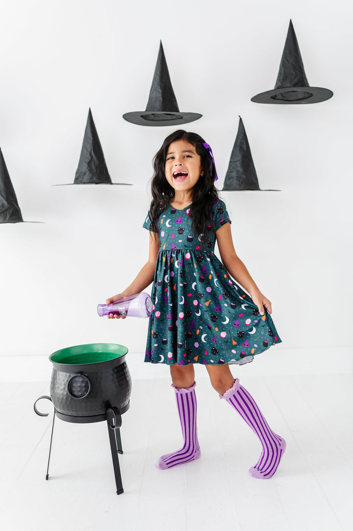 little girl surrounded by floating witch hats while pouring potion into cauldron wearing teal witchy themed twirl dress paired with two-tone purple striped knee socks