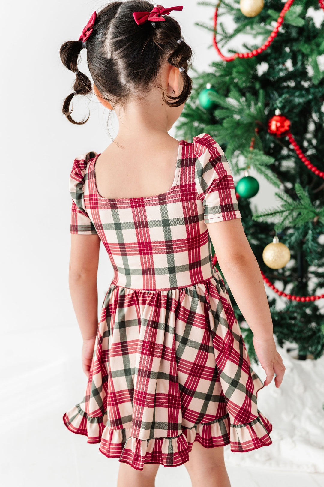 A young girl with her hair in two pigtails wearing a red, green, and beige plaid dress, photographed from behind as she stands next to a decorated Christmas tree.