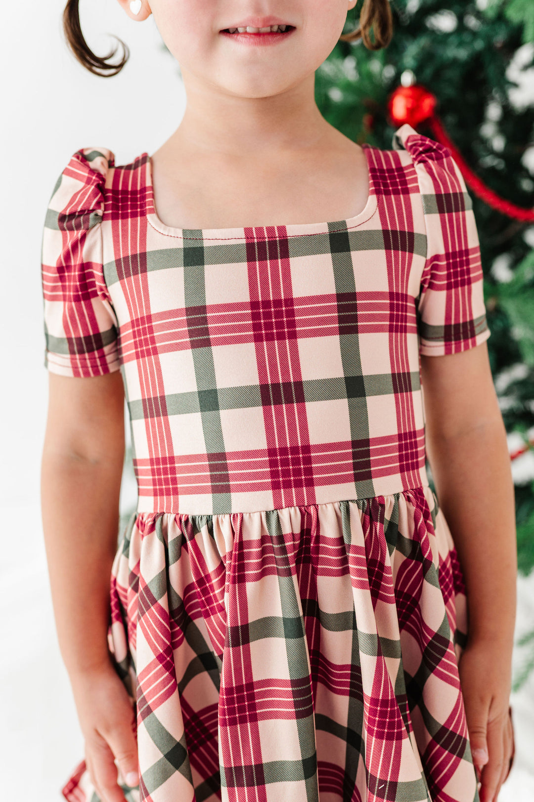 A young girl wearing a short-sleeve plaid dress with puff sleeves, a square neckline, and a gathered skirt. The dress features a red, green, and cream plaid pattern.