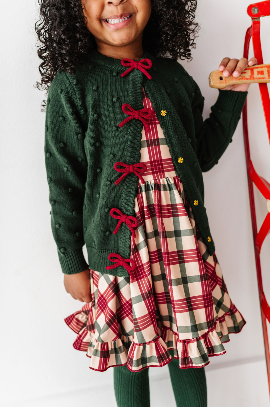 A young child wearing a dark green knit cardigan with red bow details over a red, green, and beige plaid dress, standing next to a red ladder against a white wall.