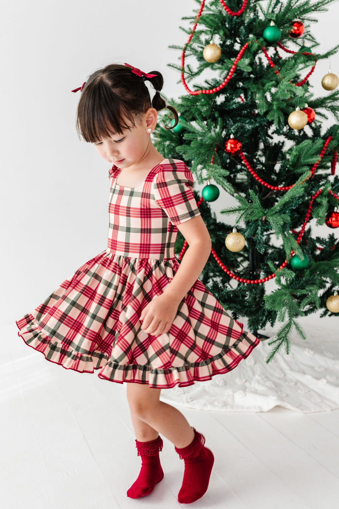 A young girl wearing a red and beige plaid short-sleeve dress and red socks stands in front of a decorated Christmas tree, looking down.