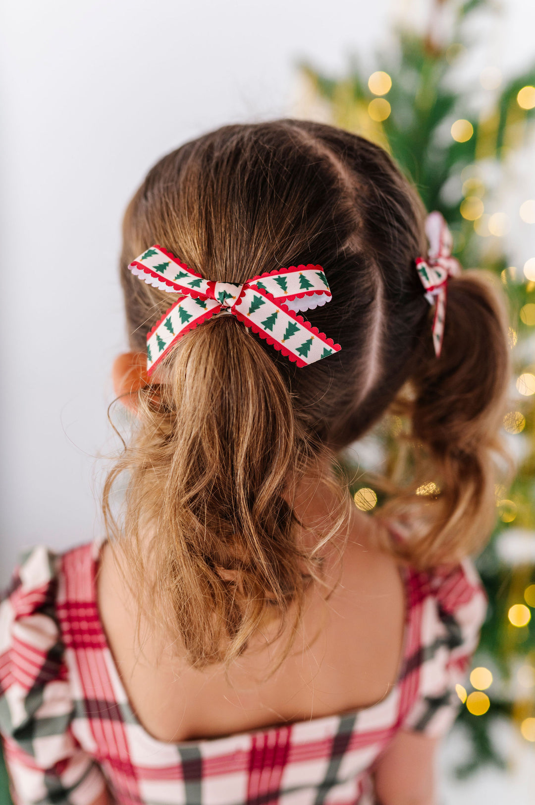 little girl with pigtails and xmas tree print ribbon bows in each ponytail wearing festive plaid dress