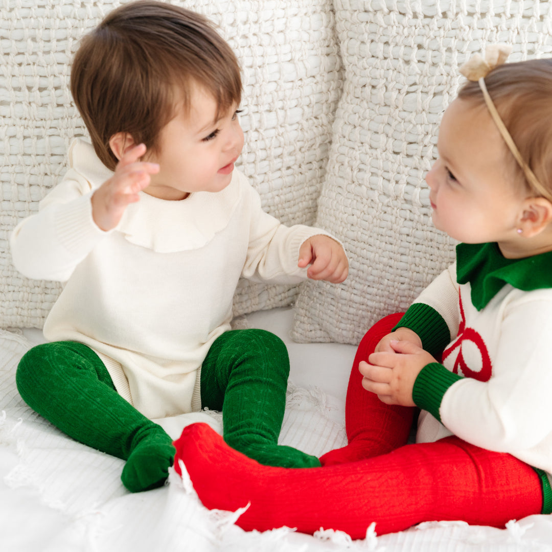 two baby girls sitting on bed wearing christmas sweater rompers with red and green baby tights