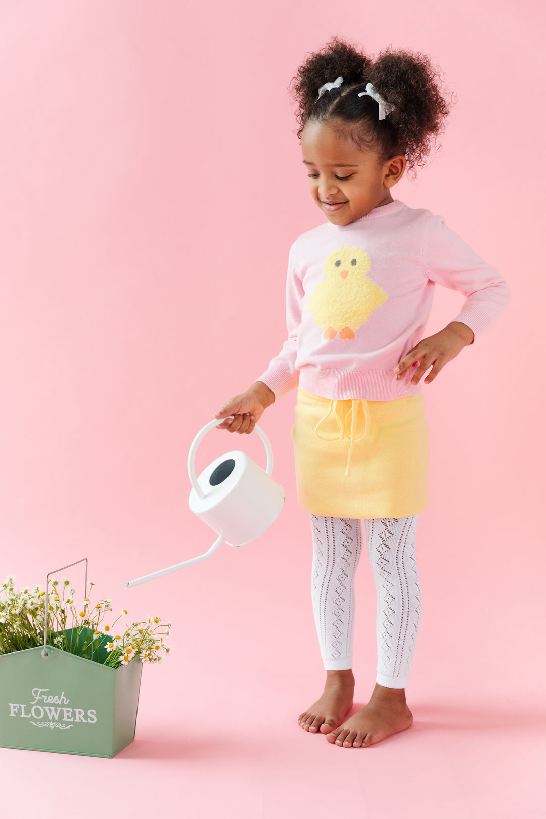 little girl watering flowers in pink fuzzy chick sweater with matching pastel yellow skirt and white footless pointelle tights