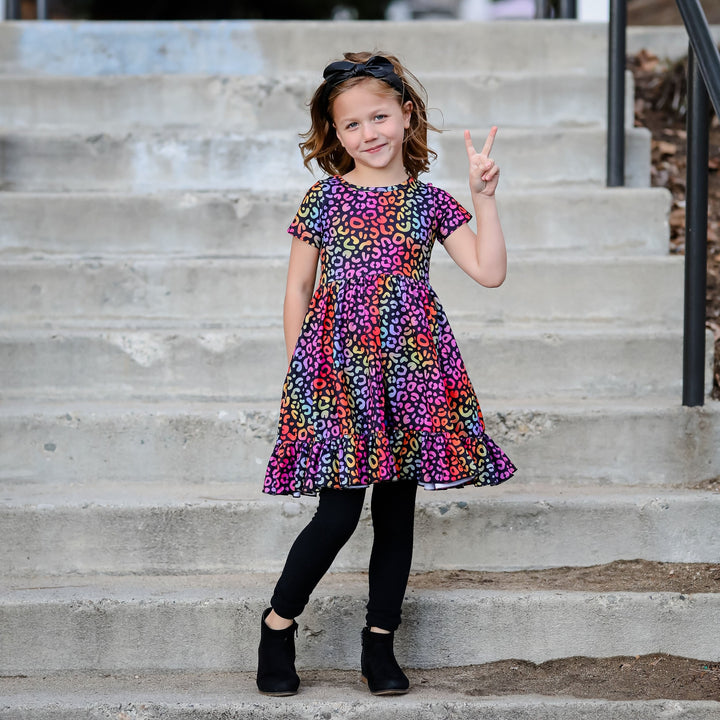 little girl giving peace sign in rainbow leopard print dress and black cable knit leggings