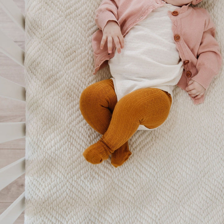 baby lounging in crib wearing white onesie and mustard cable knit tights underneath
